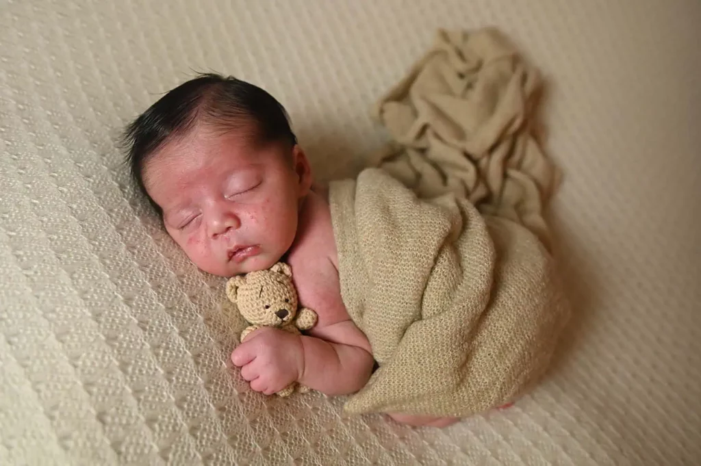 A sleeping baby lies on a textured beige blanket, wrapped snugly in a light brown knit wrap. The baby holds a small crocheted teddy bear close to their chest, creating a serene and cozy atmosphere that feels like home. - Image by Kristin Milito Photography, Chicago IL.