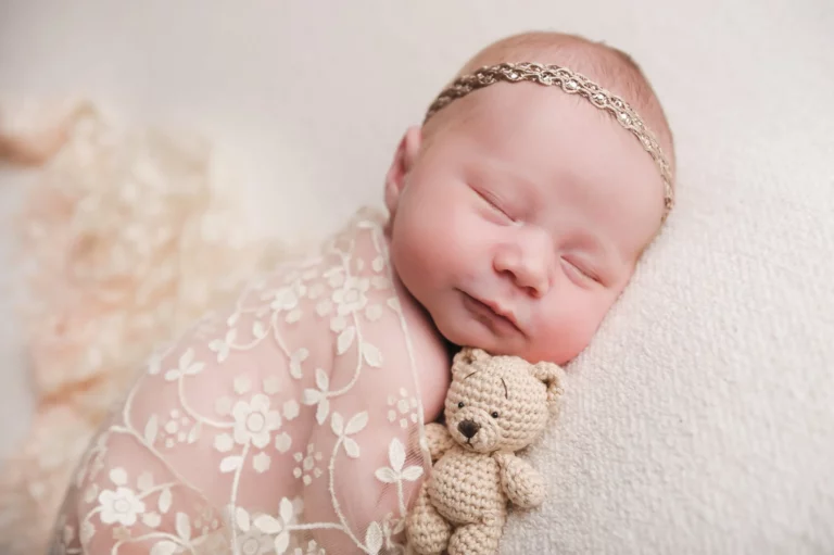 A peacefully sleeping baby, wrapped in a delicate, flower-patterned pink blanket, rests on its side. The baby wears a thin, decorative headband and gently holds a small, knitted teddy bear. The soft white background enhances the serene and tender scene, evoking the warmth of home. - Image by Kristin Milito Photography, Chicago IL.