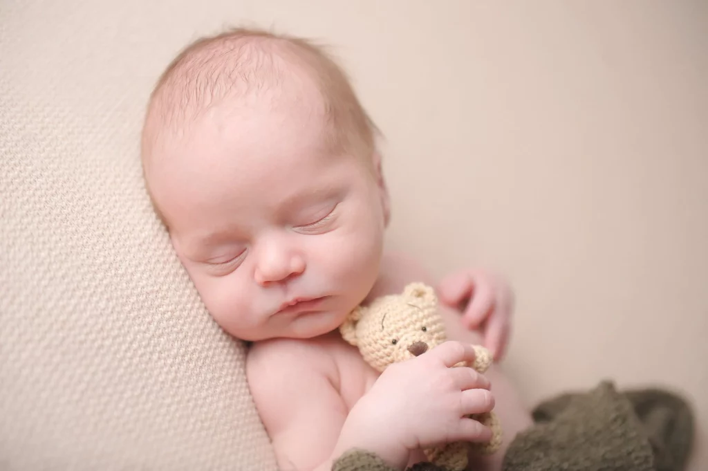A sleeping baby, lying on a beige textured surface, gently cradles a small knitted teddy bear. The baby's eyes are closed and they appear peaceful with a slight smile. The image is soft and tranquil, conveying a sense of calm and innocence. - Image by Kristin Milito Photography, Chicago IL.