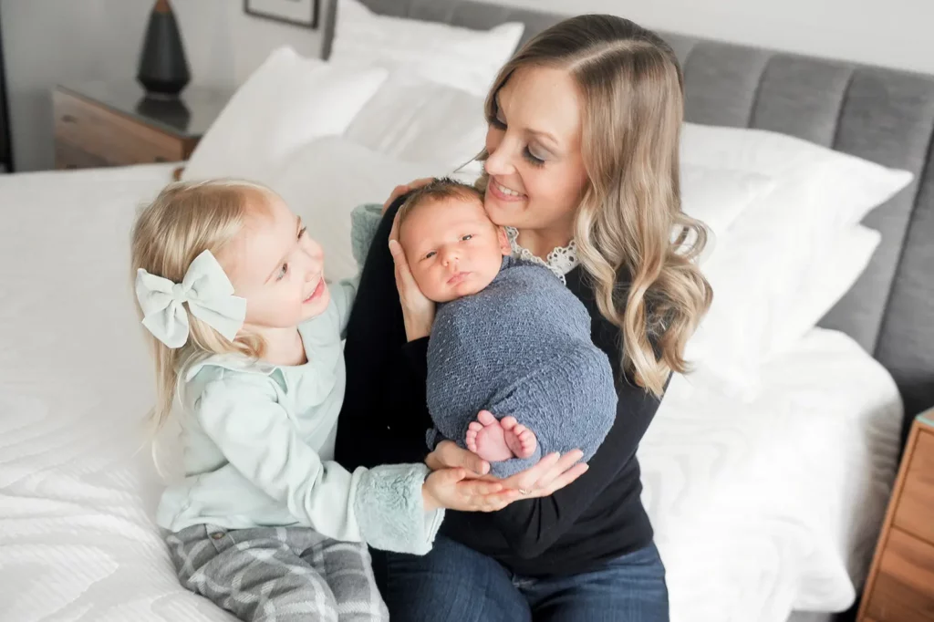 A woman sits on a bed holding a swaddled newborn while smiling at a young girl with a large bow in her hair. Captured by a Chicago Lifestyle Photographer, the joyful family is surrounded by soft white bedding. Kristin Milito Photography - Newborn & Maternity Photographer - Chicago IL