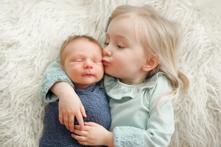 A young child with long blonde hair kisses a newborn baby wrapped in a blue blanket while cuddling together on a soft, white, fluffy surface, perfectly capturing the tender moments by a Chicago lifestyle photographer. Kristin Milito Photography - Newborn & Maternity Photographer - Chicago IL