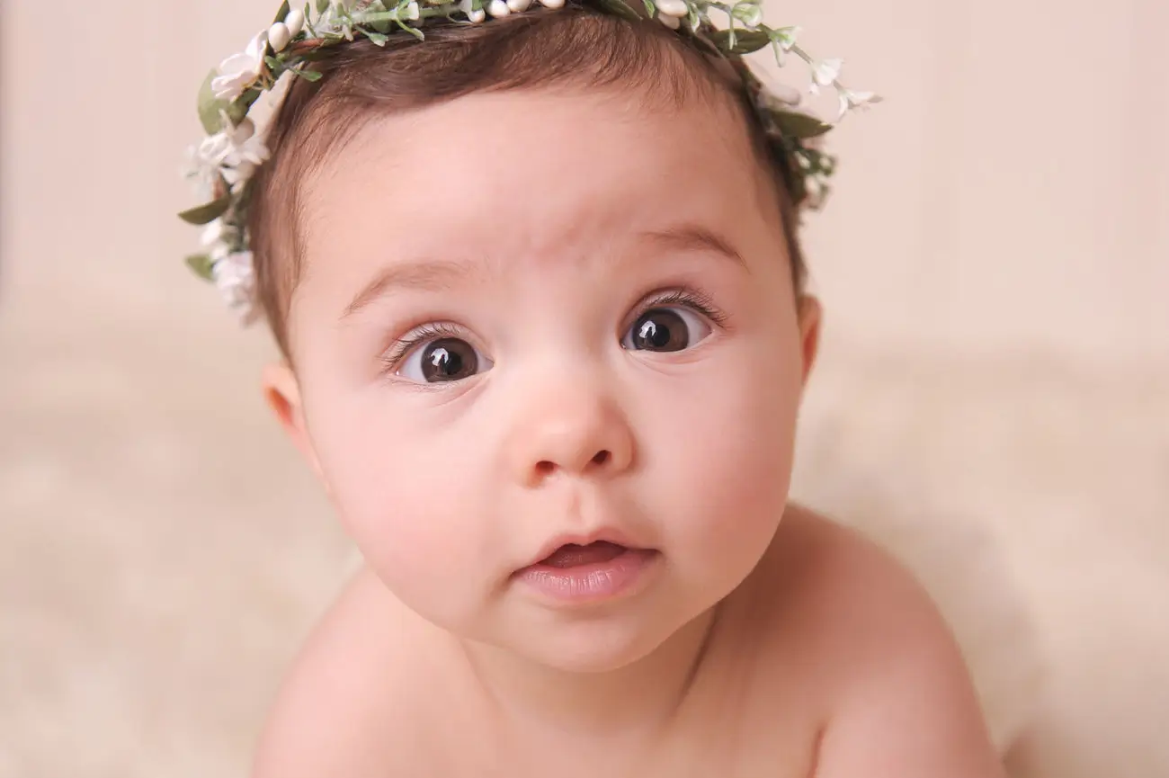 A close-up of a baby with big brown eyes and light skin, looking curiously at the camera. Captured by a Chicago Newborn Photographer, the baby wears a delicate floral headband against a soft, neutral background. Kristin Milito Photography - Newborn & Maternity Photographer - Chicago IL