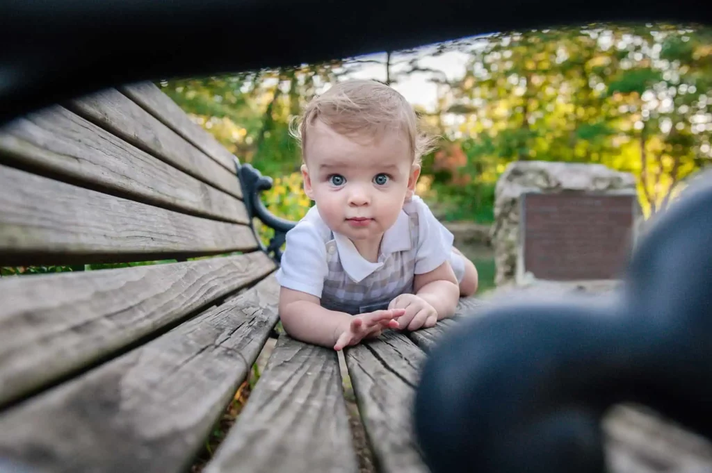A baby in light-colored overalls crawls on a wooden park bench, looking curiously at the camera