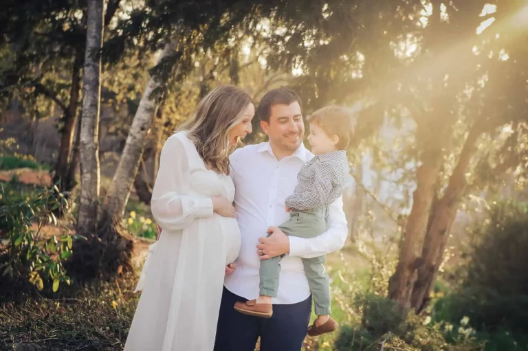 A pregnant woman in a white dress stands outdoors with a man holding a small child. They are smiling at each other, surrounded by trees and sunlight streaming through the branches. Kristin Milito Photography - Newborn & Maternity Photographer - Chicago IL