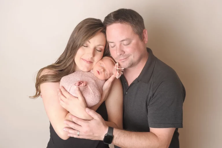 A woman and man gently cradle a sleeping newborn wrapped in a pink blanket. All three have their eyes closed, sharing a tender, peaceful moment—beautifully captured by a Chicago Family Photographer against a light, neutral background