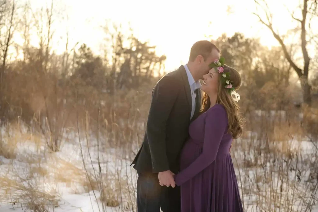A couple stands in a snowy field at sunset, holding hands. The man is wearing a suit and gently kissing the woman's forehead. The woman, wearing a purple dress and a floral crown, smiles warmly, her pregnant belly visible. The scene is serene with barren trees in the background. - Image by Kristin Milito Photography, Chicago IL.