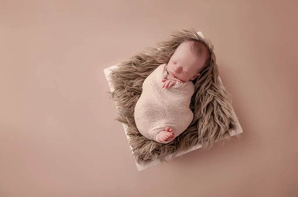 A newborn baby sleeps peacefully, swaddled in a beige blanket. The baby is lying on a fluffy grey mat atop a white square cushion. The background is a warm, solid light brown, creating a cozy atmosphere. The baby's feet and delicate face are visible and relaxed. - Image by Kristin Milito Photography, Chicago IL.