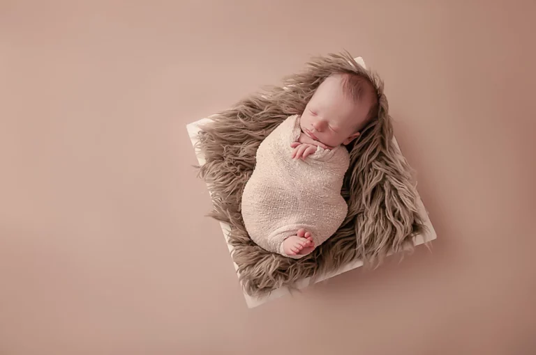 A newborn baby sleeps peacefully, swaddled in a beige blanket. The baby is lying on a fluffy grey mat atop a white square cushion. The background is a warm, solid light brown, creating a cozy atmosphere. The baby's feet and delicate face are visible and relaxed.