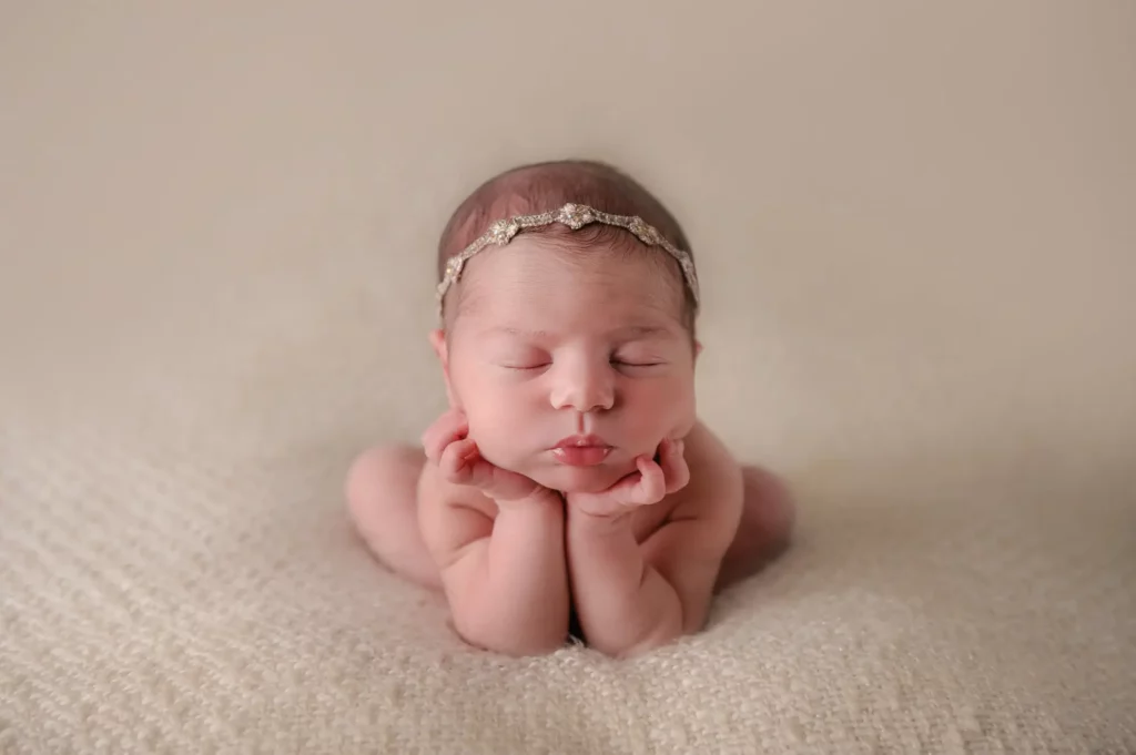 A Chicago newborn photographer captured a serene and peaceful scene of a newborn baby, wearing a delicate headband, resting on a soft, neutral-toned blanket. The baby is posed with their chin on their hands, eyes closed, and legs tucked under their body. - Image by Kristin Milito Photography, Chicago IL.