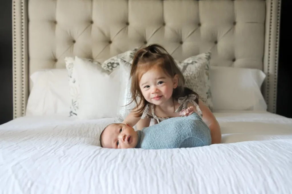 A young girl smiles while kneeling on a bed behind a swaddled newborn baby, both on a white comforter with plush pillows behind them, captured by a Lifestyle Photographer in Chicago. Kristin Milito Photography - Newborn & Maternity Photographer - Chicago IL