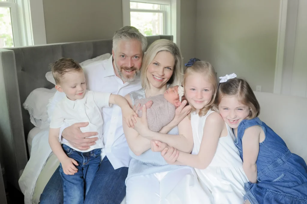 A smiling family of five, photographed by a Chicago Lifestyle Photographer, sits closely together on a bed in a bright, softly lit room with their newborn baby, two young daughters, and young son. Kristin Milito Photography - Newborn & Maternity Photographer - Chicago IL