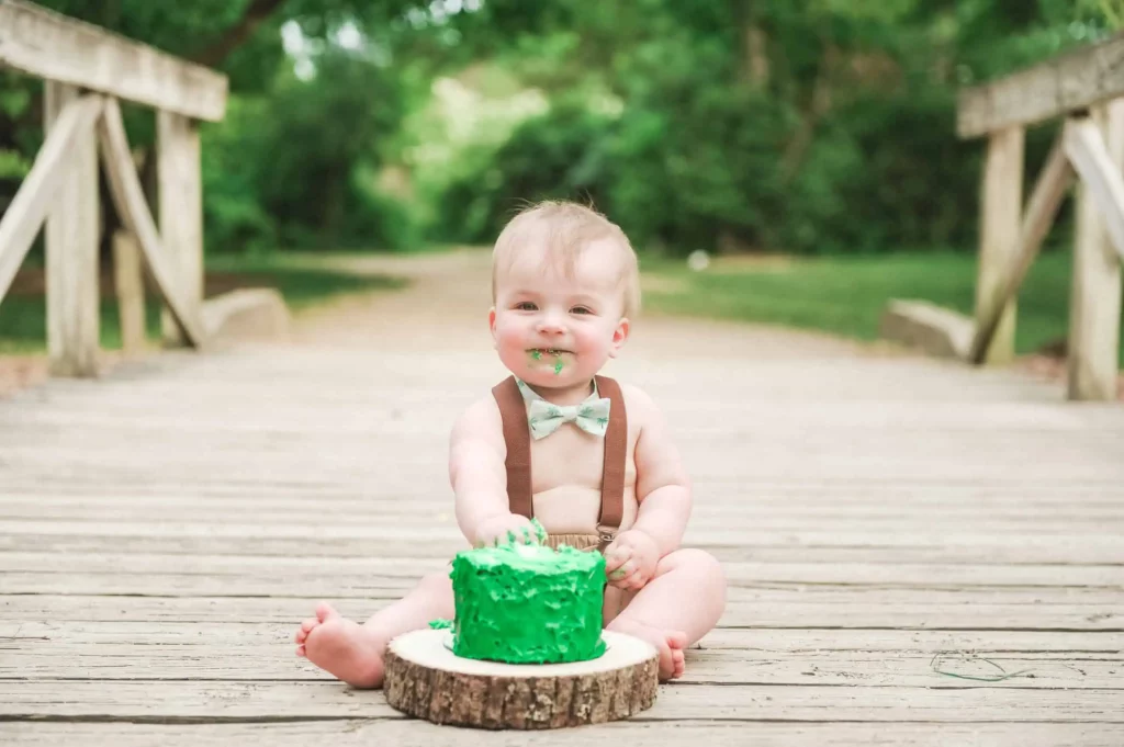 A baby wearing suspenders and a bow tie sits on a wooden bridge outdoors, smiling with green frosting on their face and