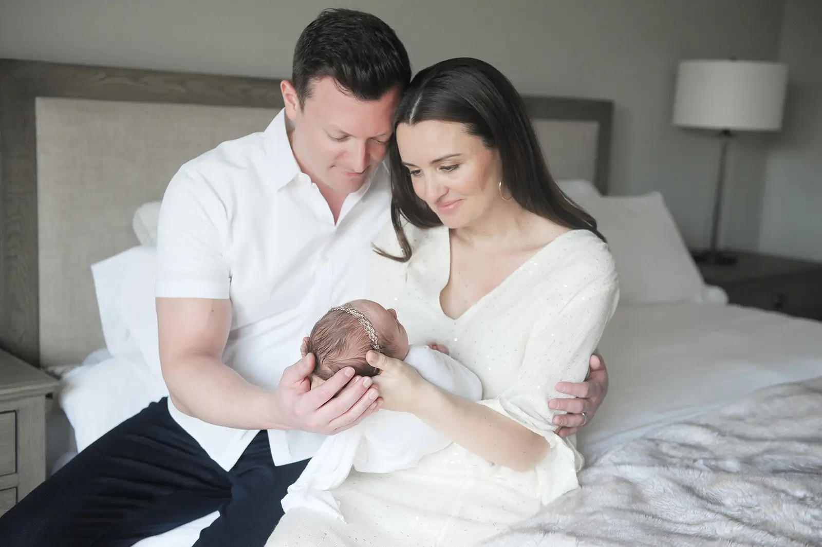 A couple sits closely together on a bed, lovingly gazing at a newborn baby wrapped in a white blanket, whom the mother is cradling in her arms. The scene is soft and intimate, with neutral colors in the room. Kristin Milito Photography - Newborn & Maternity Photographer - Chicago IL