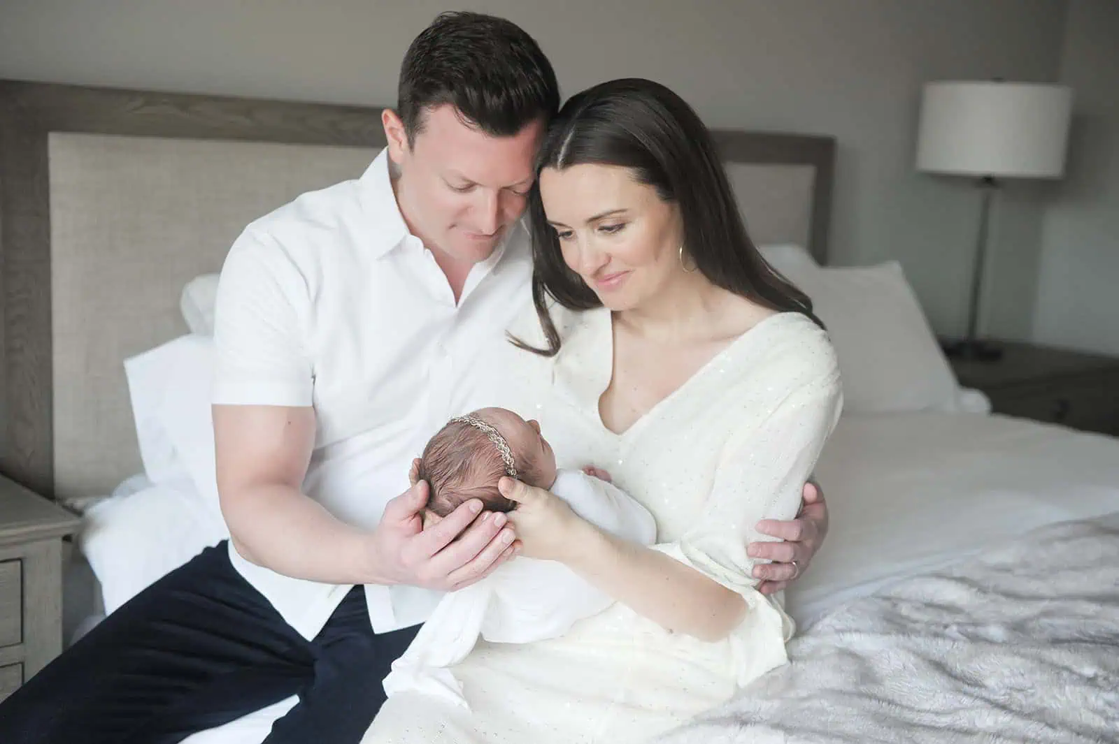 A couple sits on a bed, lovingly gazing at their newborn baby. The man gently cradles the baby's head while the woman supports their body. They are dressed in white, and the room is softly lit, reflecting a peaceful and intimate moment. - Image by Kristin Milito Photography, Chicago IL.