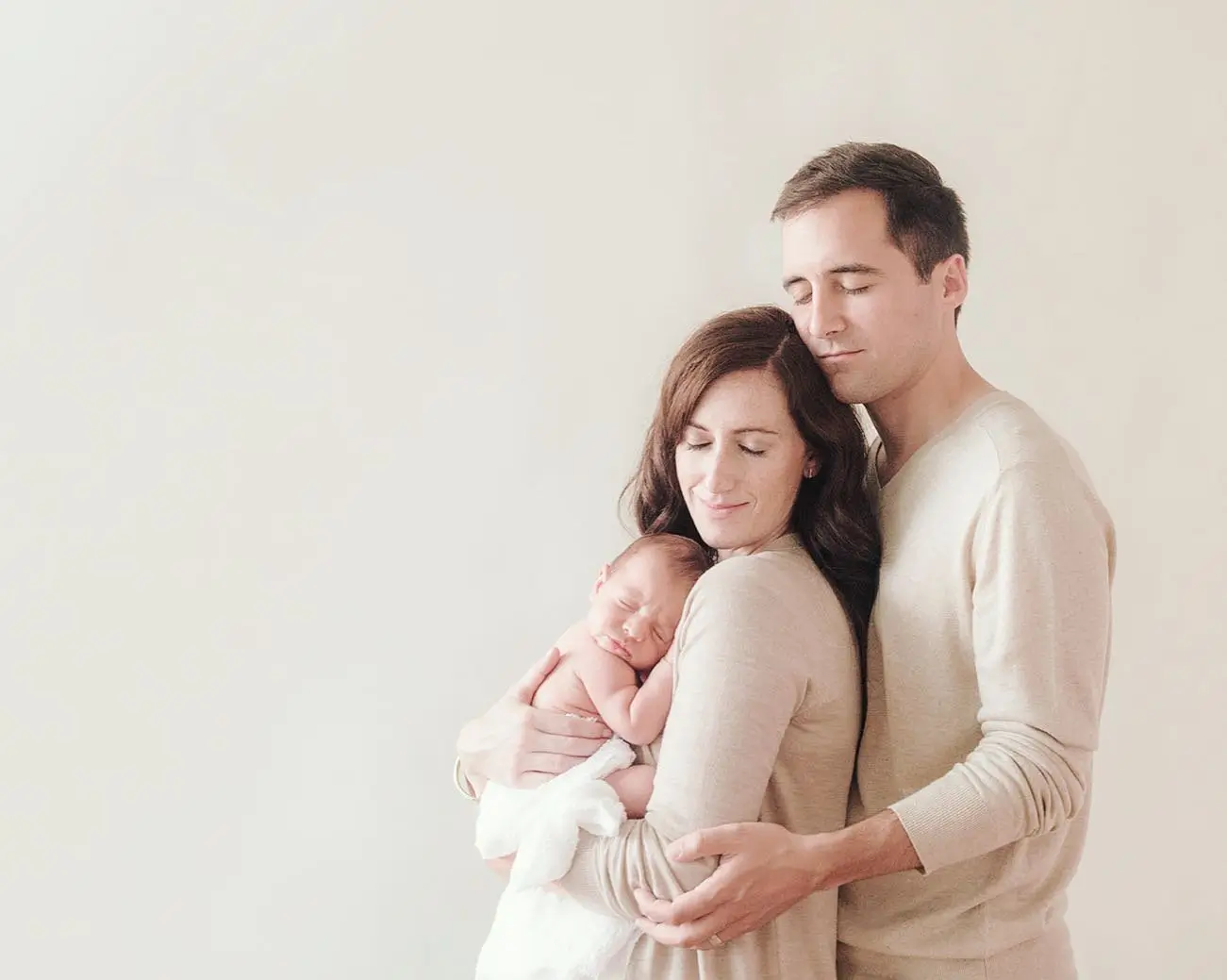 A man and woman gently embrace while holding a sleeping baby. The woman, with shoulder-length brown hair, cradles the infant against her chest, and the man, with short dark hair, wraps his arms around them both. Their matching light-colored clothing resembles a harmonious investment in family unity and love. - Image by Kristin Milito Photography, Chicago IL.