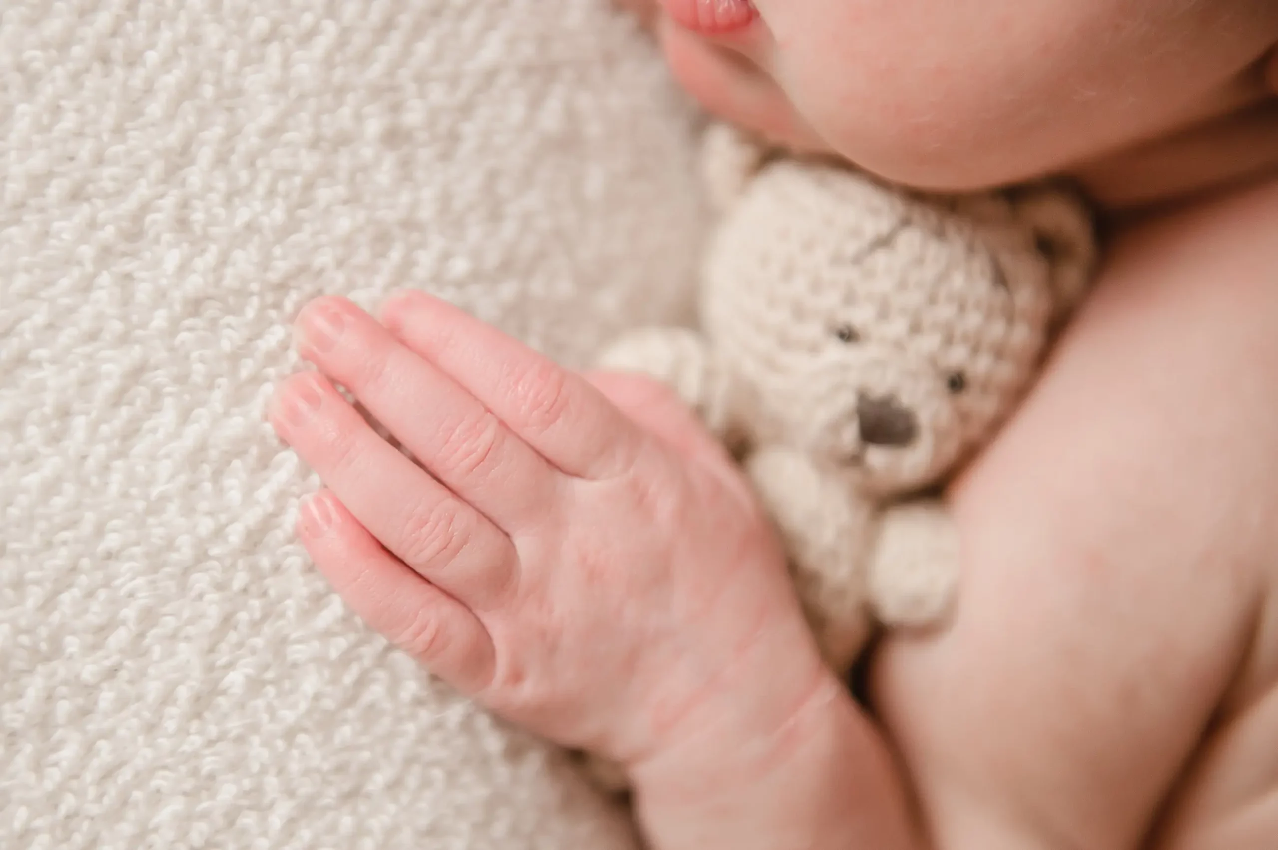 A close-up by a Chicago Newborn Photographer of a baby cuddling a small knitted teddy bear, with the baby’s hand resting gently on a soft, textured blanket. Only the lower part of the baby’s face, hand, and toy are visible. Kristin Milito Photography - Newborn & Maternity Photographer - Chicago IL