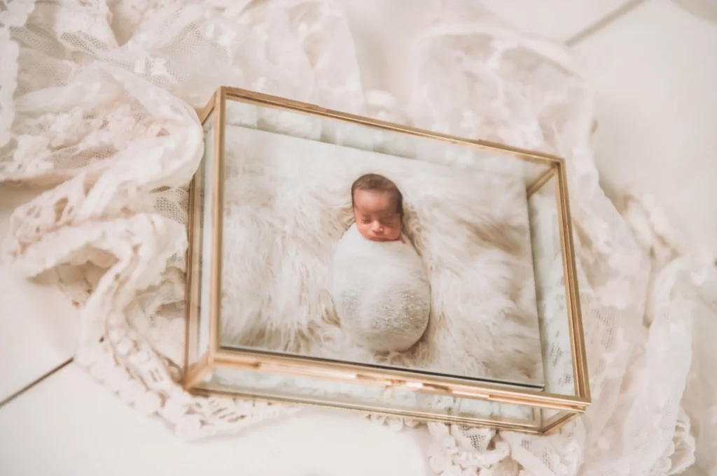 A framed photograph of a swaddled newborn baby lies on a soft, white, lace fabric background, creating a delicate and