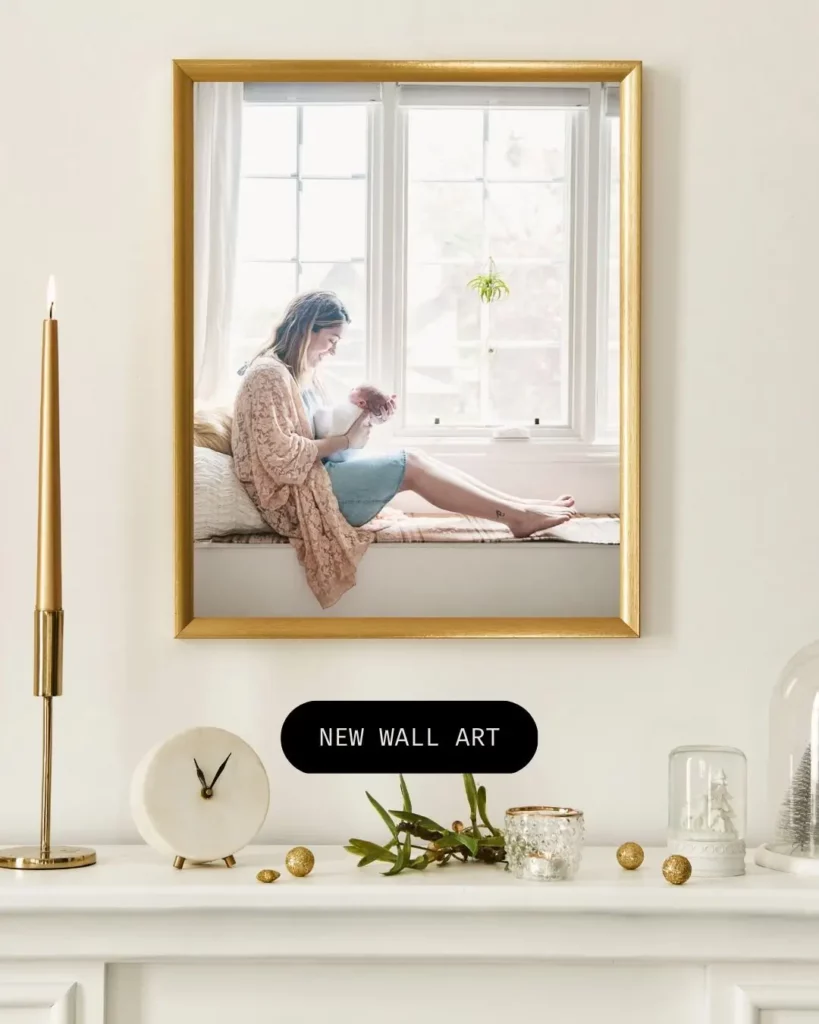 A framed photo of a woman sitting by a bright window hangs on a white wall above a shelf decorated with a gold candle, a clock, greenery, and small ornaments. Text below the frame reads "NEW WALL ART. Kristin Milito Photography - Newborn & Maternity Photographer - Chicago IL
