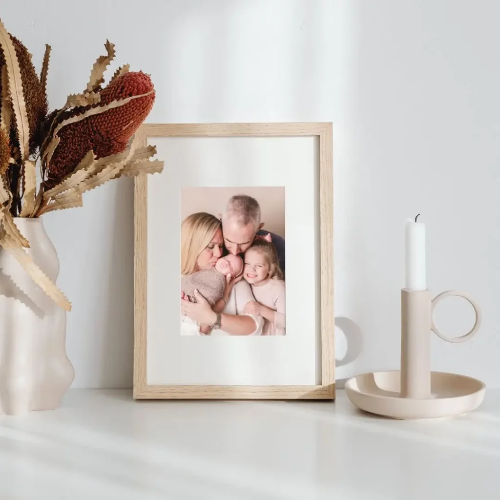 A framed photo of a smiling family with two adults and two children sits on a white surface. Next to it are a beige vase with dried flowers and a beige candle holder with a white candle. Sunlight casts soft shadows. Kristin Milito Photography - Newborn & Maternity Photographer - Chicago IL