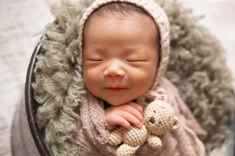 A newborn baby wrapped in a soft, light brown blanket sleeps peacefully, smiling with eyes closed, holding a small knitted teddy bear. The baby is nestled against a fluffy, gray cushion inside a dark basket, wearing a matching knitted bonnet. - Image by Kristin Milito Photography, Chicago IL.