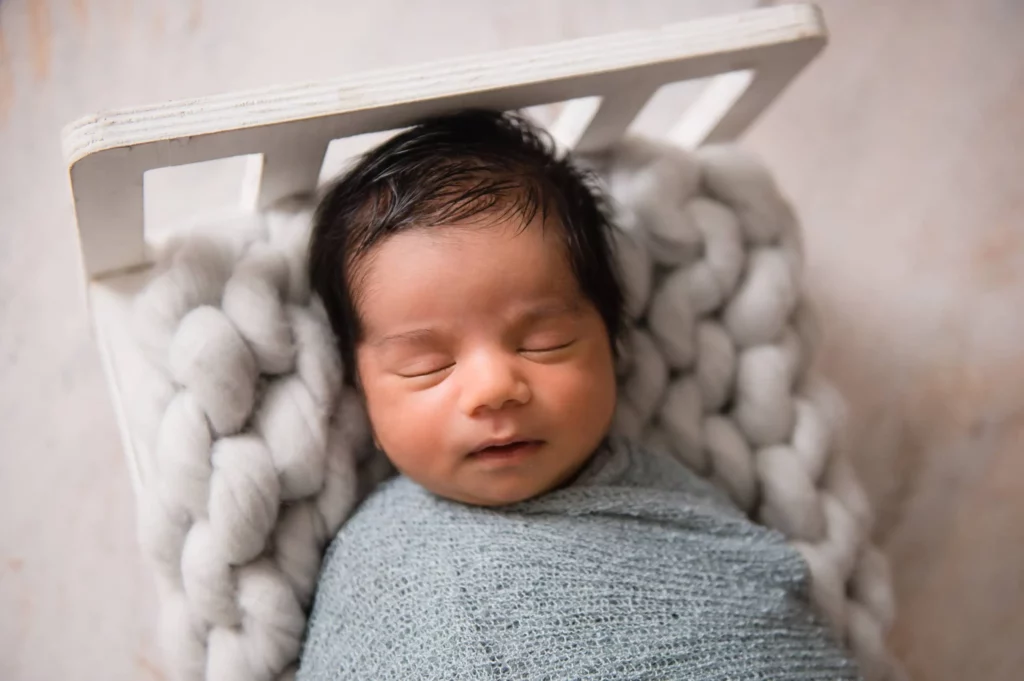 A newborn baby with dark hair sleeps peacefully, wrapped in a light gray blanket. The baby is resting on a white, knitted cushion inside a small white bed. The overall atmosphere appears serene and cozy. - Image by Kristin Milito Photography, Chicago IL.