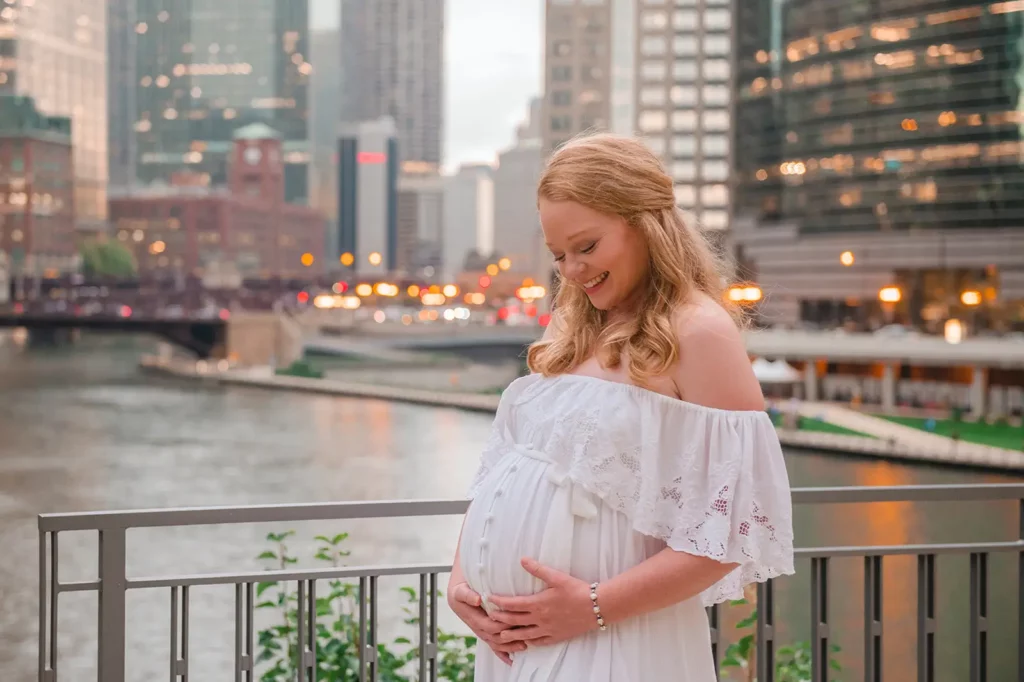 A pregnant woman in a white off-shoulder dress stands by a riverfront railing, smiling and gently cradling her belly. Tall buildings and lights create the perfect backdrop captured by a Chicago Maternity Photographer. Kristin Milito Photography - Newborn & Maternity Photographer - Chicago IL
