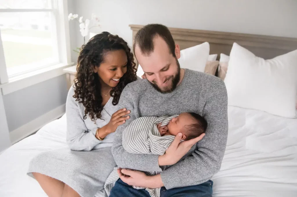 A smiling couple sits on a bed; the man cradles a newborn baby wrapped in a striped blanket while the woman lovingly touches his shoulder. The bright, cozy room with white bedding captures this heartfelt moment by a Chicago Family Photographer. Kristin Milito Photography - Newborn & Maternity Photographer - Chicago IL