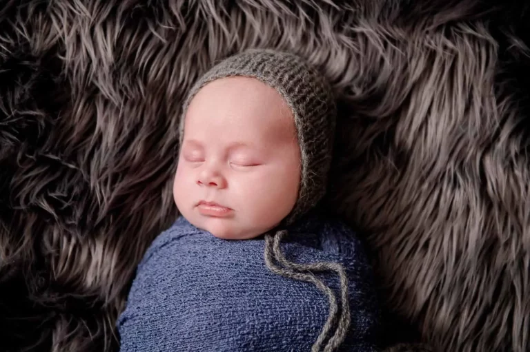 A sleeping baby, bundled in a blue wrap and wearing a knitted green bonnet, lies peacefully on a soft, furry grey blanket. - Image by Kristin Milito Photography, Chicago IL.