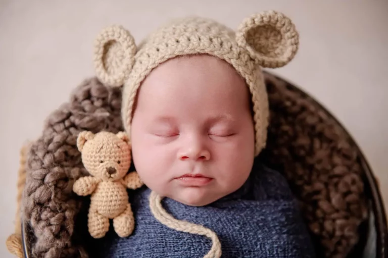 A sleeping baby, swaddled in a blue blanket, wears a crocheted hat with bear ears. The baby is nestled in a knitted blanket and cuddles a small crocheted teddy bear. The background is soft and neutral, providing a cozy feel to the image. - Image by Kristin Milito Photography, Chicago IL.