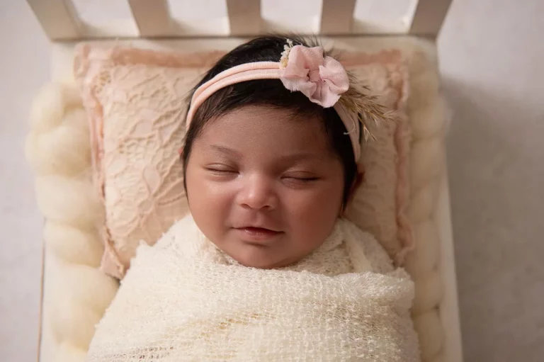 A newborn baby wrapped in a soft, cream-colored blanket lies peacefully on a cushioned surface. The baby has dark hair adorned with a beige headband featuring a delicate pink flower and feather accent. The baby’s eyes are closed, with a faint smile on its face. - Image by Kristin Milito Photography, Chicago IL.