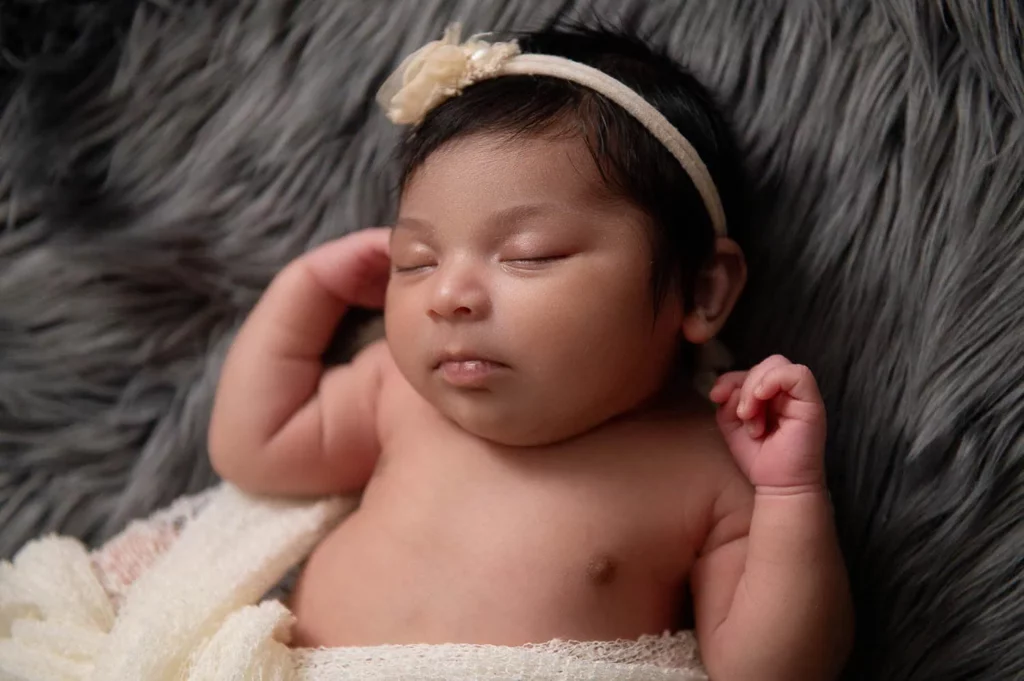 A sleeping newborn baby lies on a fuzzy gray blanket. The baby has dark hair and is wearing a cream-colored headband with a small flower. The baby is also wrapped in a light cream-colored blanket around the lower body, with one arm bent and resting near the head. - Image by Kristin Milito Photography, Chicago IL.