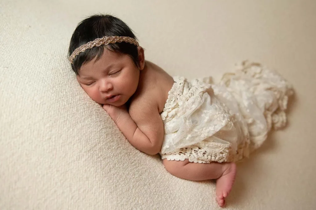 A newborn baby with dark hair is peacefully sleeping on their side, facing the camera. The baby is wearing a delicate, lace cream-colored wrap and a matching lace headband, providing a serene and soft atmosphere. The background is smooth and beige. - Image by Kristin Milito Photography, Chicago IL.