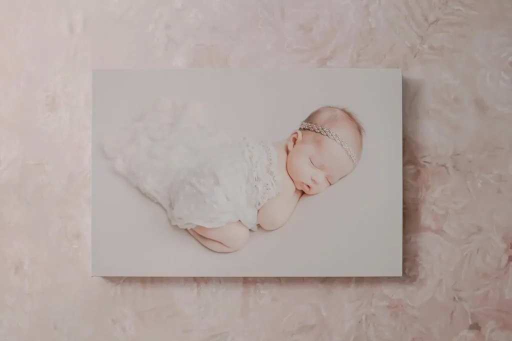 A photograph of a newborn baby sleeping on its side, dressed in a white lacy wrap and wearing a delicate headband, set against a soft, pale textured background. Kristin Milito Photography - Newborn & Maternity Photographer - Chicago IL