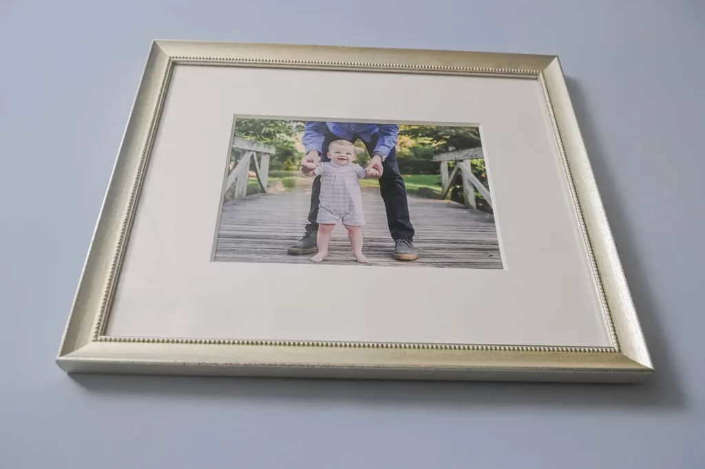 A framed photo on a light gray wall shows a smiling baby standing on a wooden bridge, supported by an adult whose hands and legs are visible behind the child. Kristin Milito Photography - Newborn & Maternity Photographer - Chicago IL