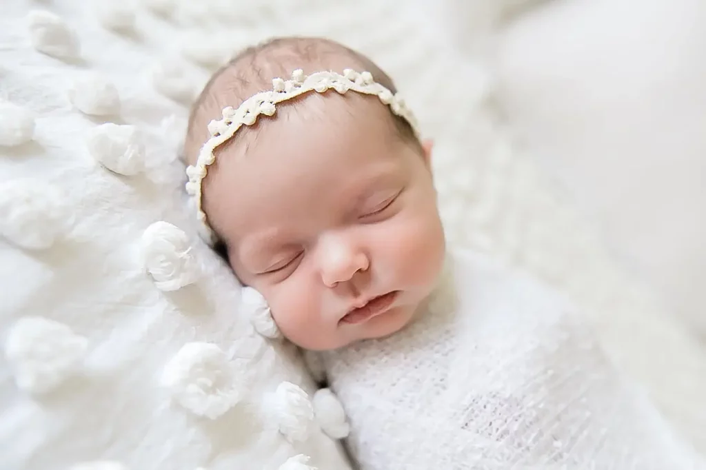 A newborn baby peacefully sleeps on a soft white blanket. The baby is swaddled in a white lace wrap and wears a delicate, white floral headband. The background is also white, creating a serene and angelic atmosphere that evokes the comfort of home. - Image by Kristin Milito Photography, Chicago IL.