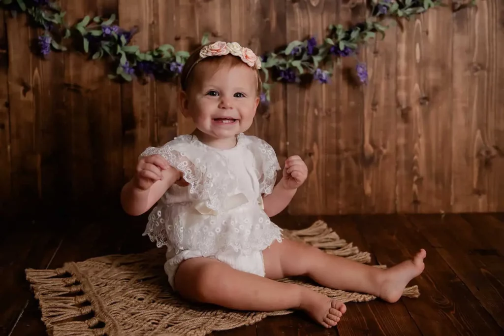 Smiling baby girl in white lace romper and flower headband against a rustic wood backdrop