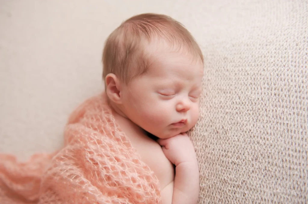 A newborn baby with light hair is peacefully sleeping on a light-textured knit blanket, bringing a sense of warmth and home. The baby is wrapped in a soft pink knitted blanket, with its tiny hands tucked under its chin and eyes gently closed. The overall scene radiates tranquility and innocence. - Image by Kristin Milito Photography, Chicago IL.