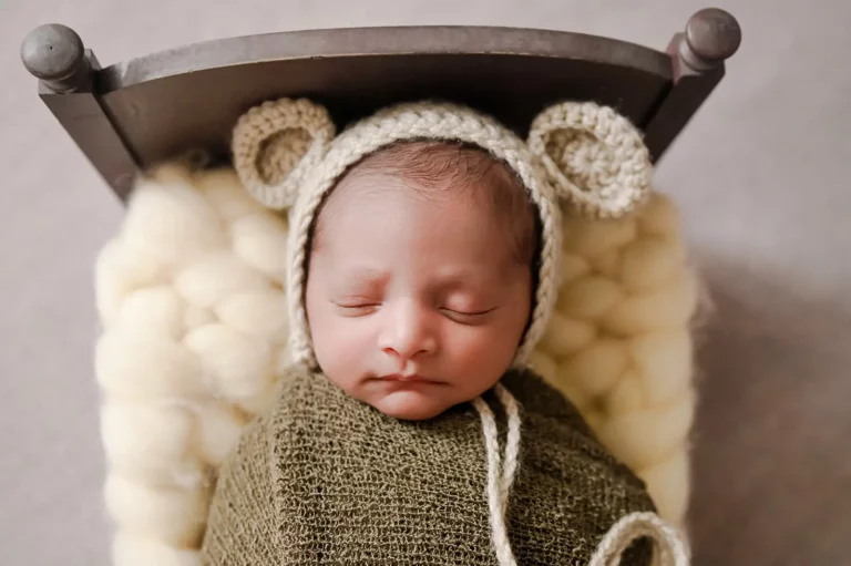 A newborn baby is sleeping peacefully at home, swaddled in a green blanket. The baby is wearing an adorable knit hat with bear ears and rests on a cushioned surface that resembles a tiny bed with a light yellow blanket underneath. - Image by Kristin Milito Photography, Chicago IL.