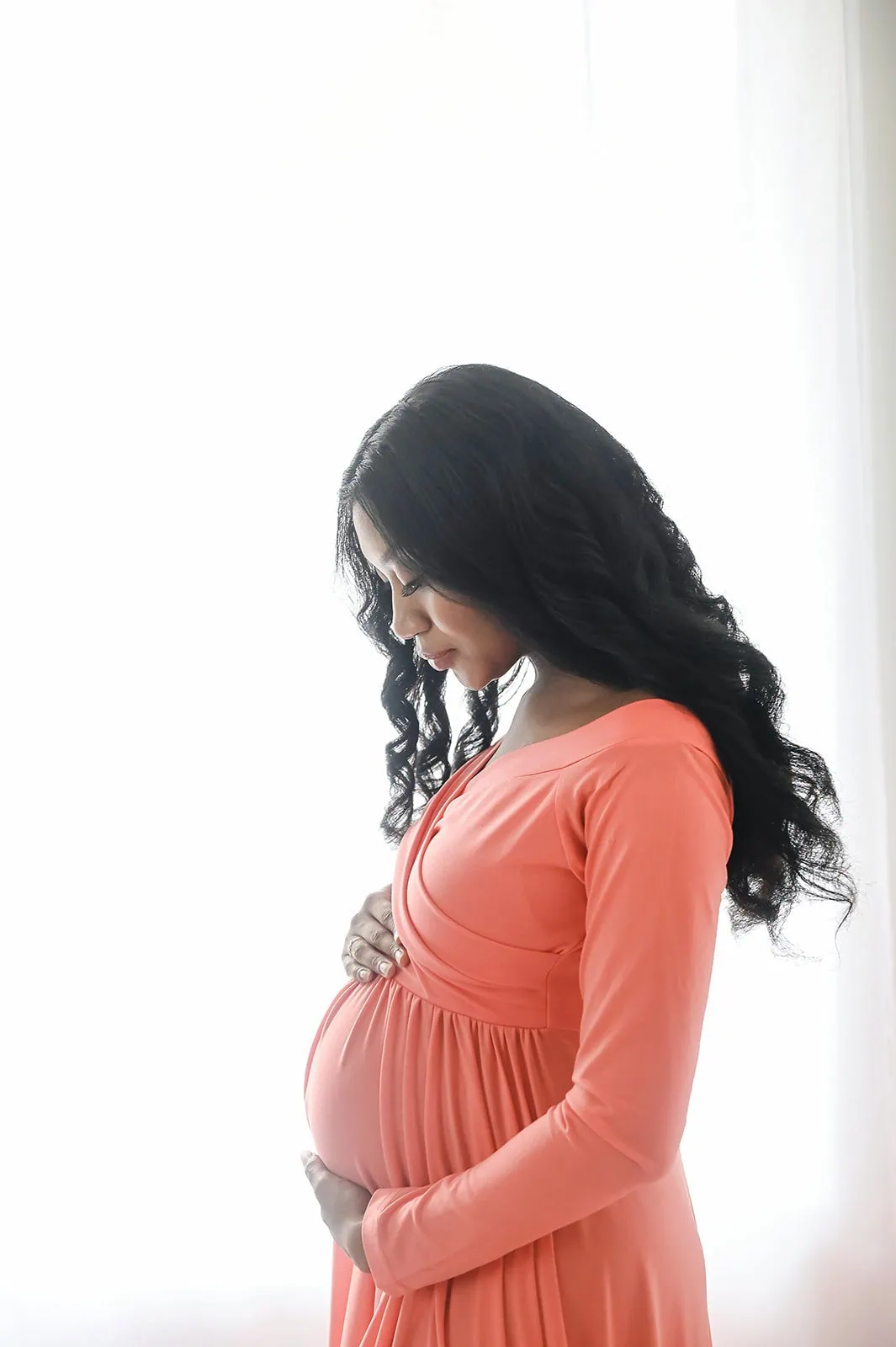 A pregnant woman with long, wavy black hair stands in profile, cradling her baby bump with both hands. She is wearing a long-sleeved coral dress and stands in front of a softly lit, white background. - Image by Kristin Milito Photography, Chicago IL.