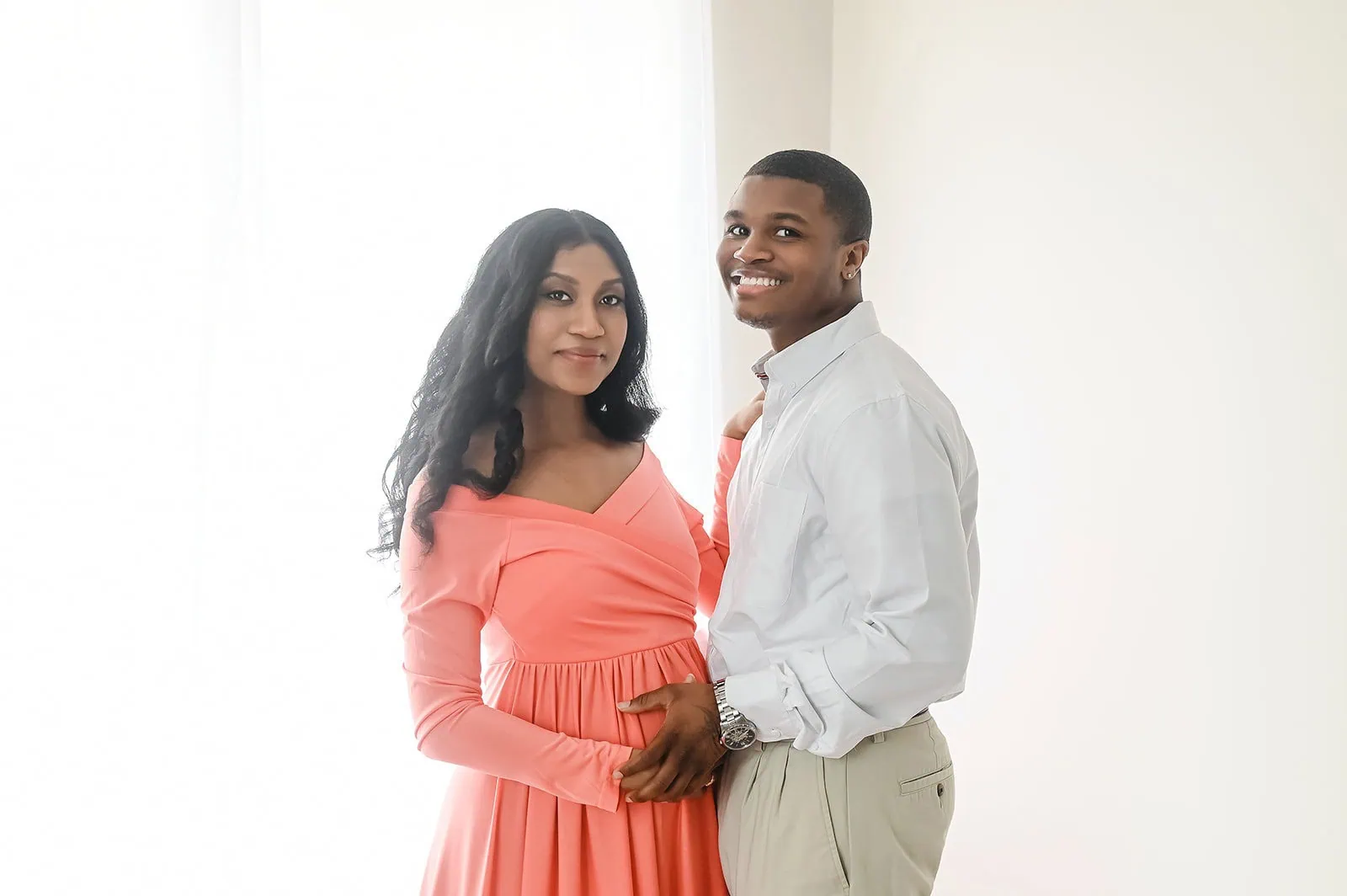 A couple stands together in front of a light background. The woman, wearing a coral dress, stands on the left with her hand on her belly. The man, in a white shirt and beige pants, stands on her right, smiling and gently placing his hand on hers. - Image by Kristin Milito Photography, Chicago IL.