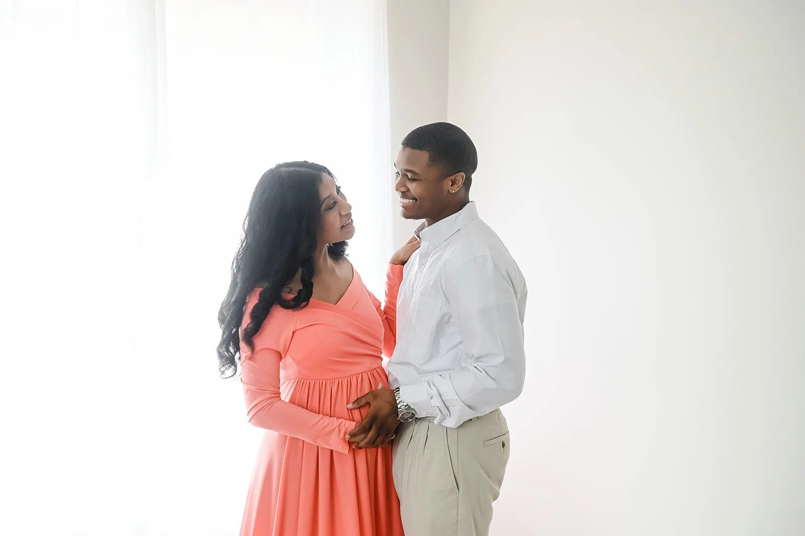 A couple stands in a brightly lit room with white walls. The woman has long, dark hair and wears a coral dress, while the man has short dark hair and wears a white shirt and light-colored pants. They are facing each other and smiling as they hold hands. - Image by Kristin Milito Photography, Chicago IL.