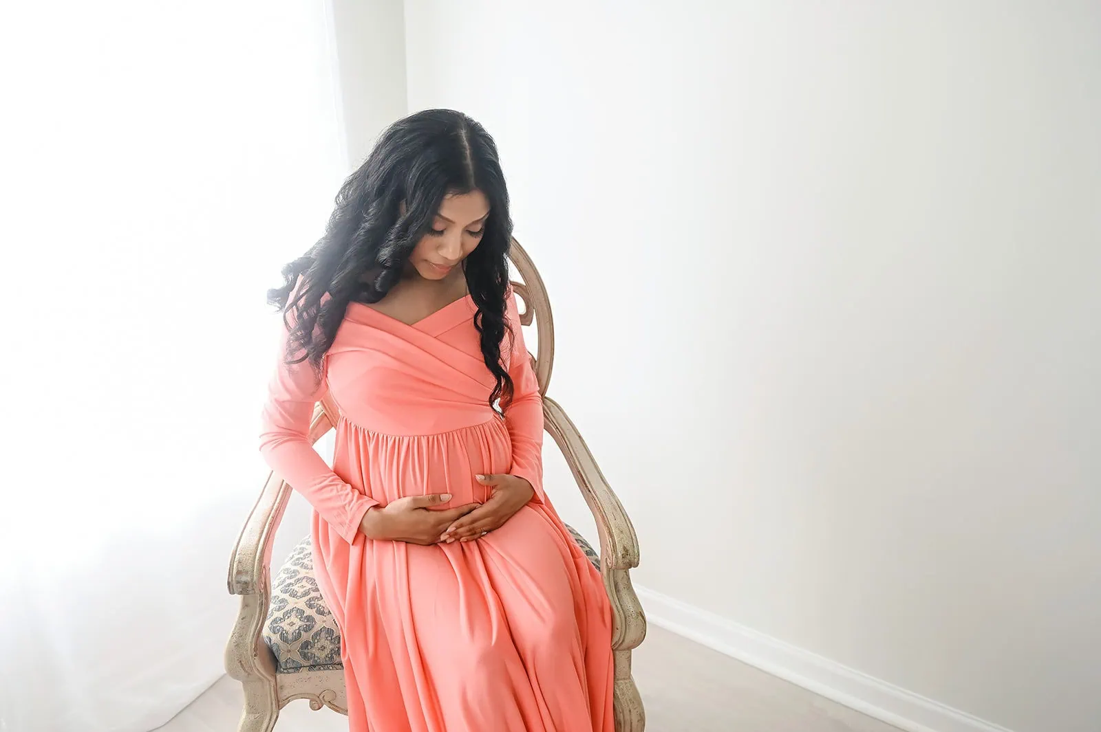 A woman with long, dark hair sits on a vintage chair by a window, bathed in soft natural light. She wears a flowing coral-colored dress and gently cradles her pregnant belly, looking down with a serene expression. The room is minimally decorated with light-colored walls. - Image by Kristin Milito Photography, Chicago IL.