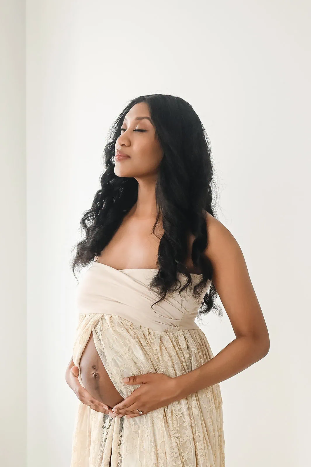 A pregnant woman with long, curly hair stands with her eyes closed, gently cradling her belly. She is wearing a strapless, cream-colored, lace dress and stands against a plain, white background, radiating a sense of calm and serenity. - Image by Kristin Milito Photography, Chicago IL.