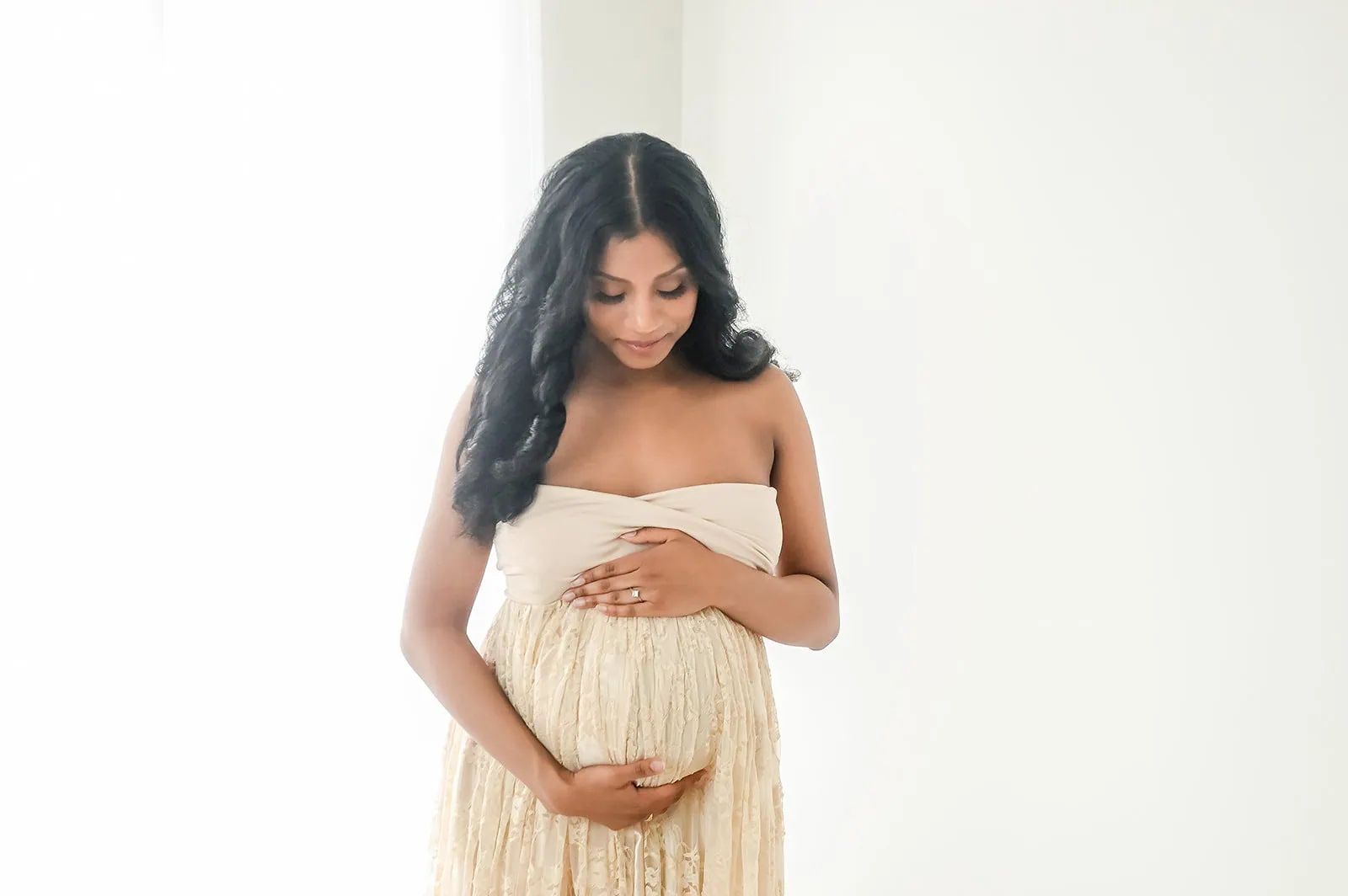 A pregnant woman with long, wavy black hair stands against a light background, gently cradling her belly with both hands. She wears a strapless cream-colored dress with a lace overlay and looks down with a serene expression. - Image by Kristin Milito Photography, Chicago IL.