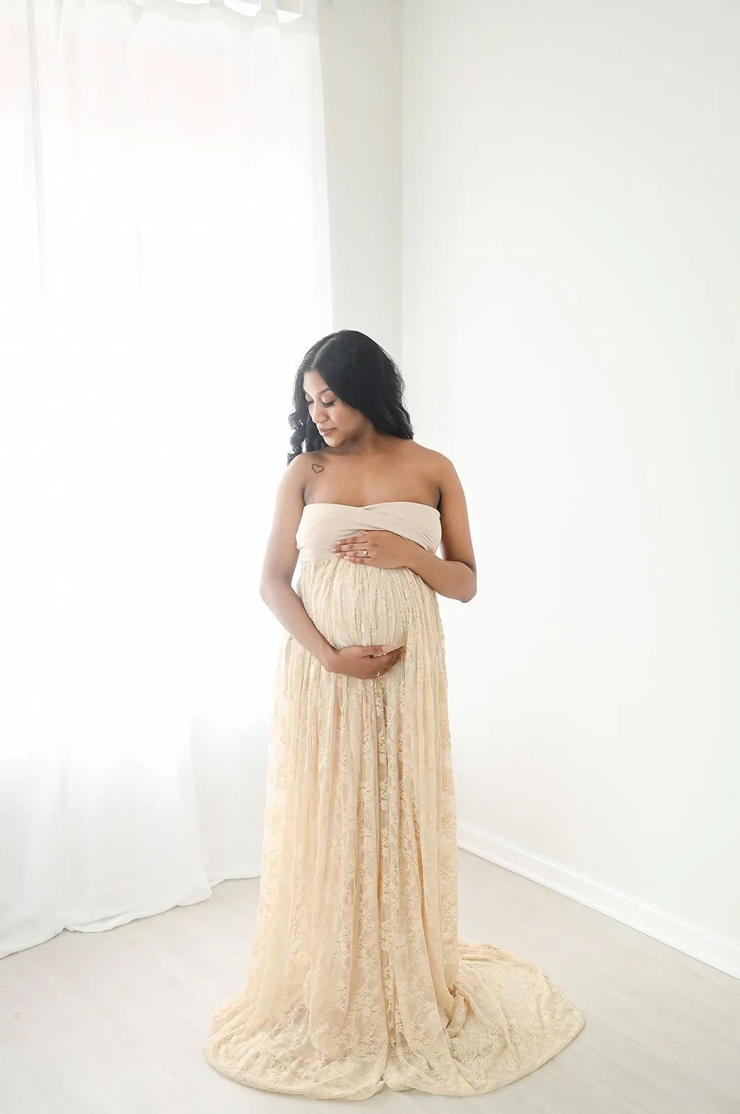 A pregnant woman stands in a bright room with sheer white curtains. She wears a strapless, flowing, lace dress and cradles her baby bump with both hands while looking down, embodying a serene and contemplative mood. - Image by Kristin Milito Photography, Chicago IL.