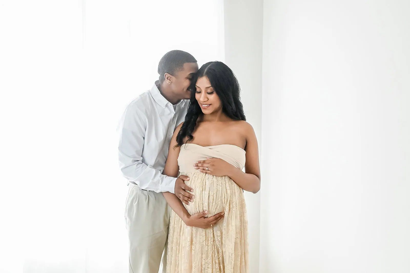 A pregnant woman in a flowing beige dress stands with her hands on her belly while a man in a light blue shirt and beige pants stands behind her, gently embracing her and resting his chin on her head. They are in a bright room with white walls and sheer curtains. - Image by Kristin Milito Photography, Chicago IL.