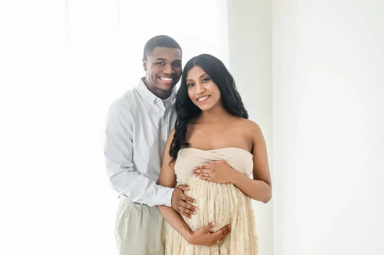 A smiling couple poses together in a bright, airy room. The woman, wearing a strapless beige dress, cradles her pregnant belly while her partner, in a light blue shirt and beige pants, stands behind her, gently placing his hands on hers. - Image by Kristin Milito Photography, Chicago IL.