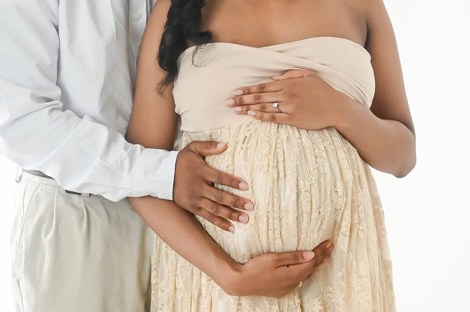 A pregnant woman in a strapless lace dress holds her belly, while a man, wearing a white shirt and light-colored pants, stands behind her, gently placing his hands over hers on her baby bump. - Image by Kristin Milito Photography, Chicago IL.