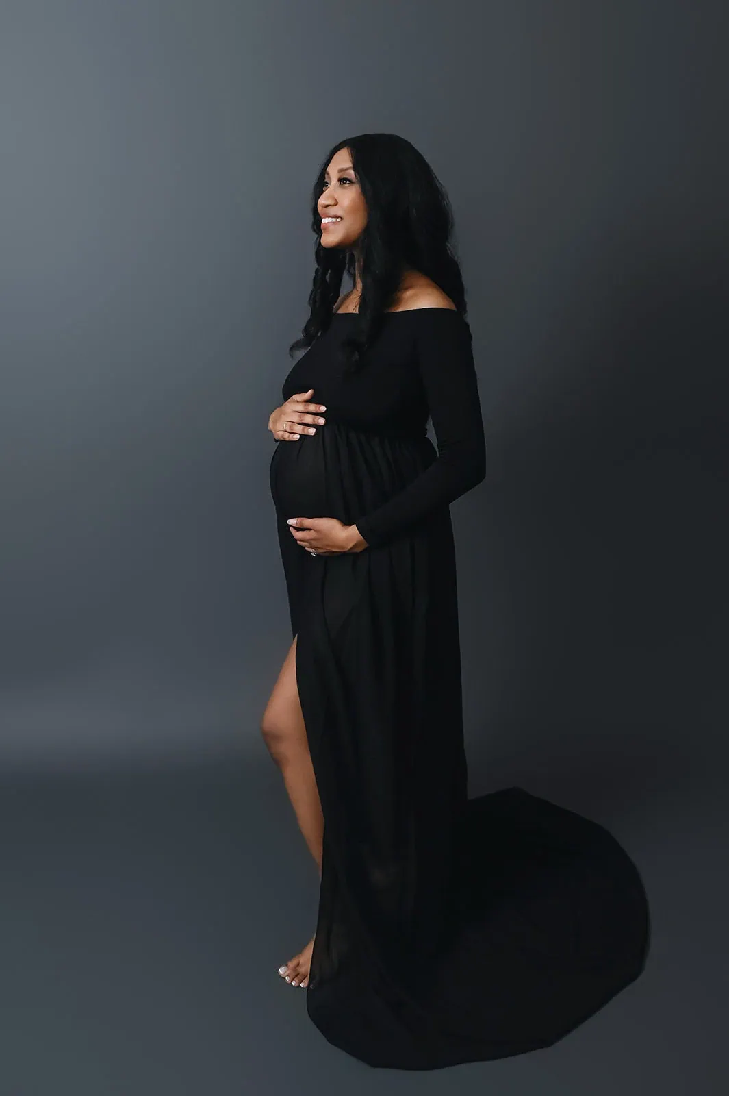 A pregnant woman wearing a long, off-the-shoulder black dress stands sideways, gently cradling her baby bump with both hands. She is looking forward with a serene expression. The background is dark and unobtrusive, keeping the focus on her. - Image by Kristin Milito Photography, Chicago IL.