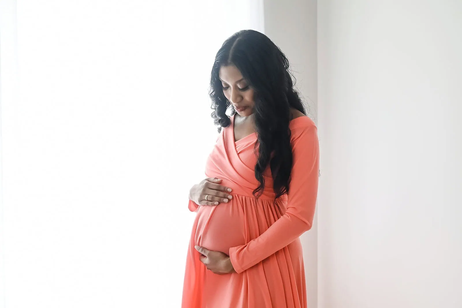 A pregnant woman with long, dark hair is gently cradling her belly with both hands. She is wearing a flowing, coral-colored dress and is standing in a softly lit, neutral-toned room, looking down lovingly at her belly. - Image by Kristin Milito Photography, Chicago IL.